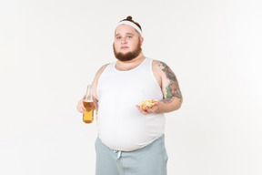 Um homem gordo em sportswear segurando uma garrafa de cerveja e batatas fritas