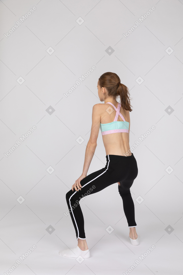 Vista posterior de tres cuartos de una jovencita en ropa deportiva en cuclillas mientras pone las manos en las piernas