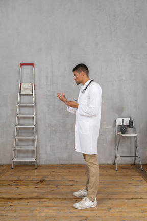 Vista lateral de un joven médico de pie en una habitación con escalera y silla explicando algo
