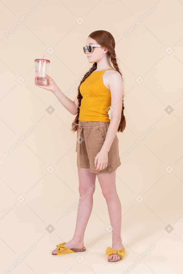 Teenager girl in sunglasses standing half sideways and holding plastic cup