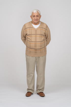 Front view of an old man in casual clothes standing with hands behind back and looking at camera