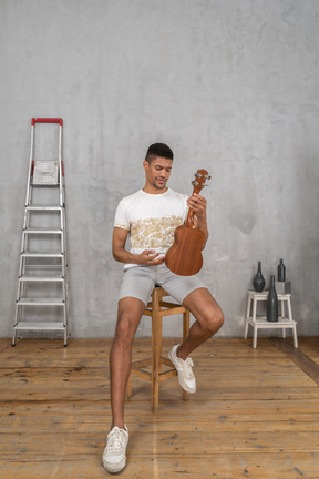 Vista frontal de un hombre en un taburete examinando un ukelele con una sonrisa