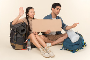 Young interracial couple showing hitchhiking sign sitting on backpacks