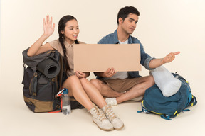 Joven pareja interracial mostrando el signo de autostop sentado en las mochilas