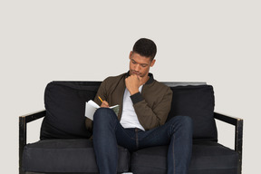Вид спереди молодого человека, сидящего на диване и делающего заметки