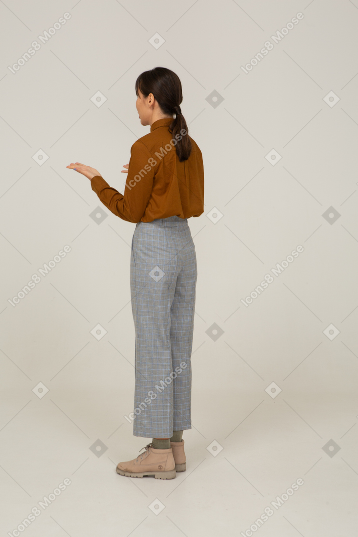 Vista posteriore di tre quarti di una giovane donna asiatica interrogativa in calzoni e camicetta che alza le mani