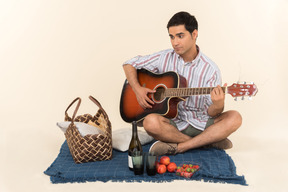 Chico caucásico joven sentado cerca de la cesta de picnic en la manta y tocando la guitarra