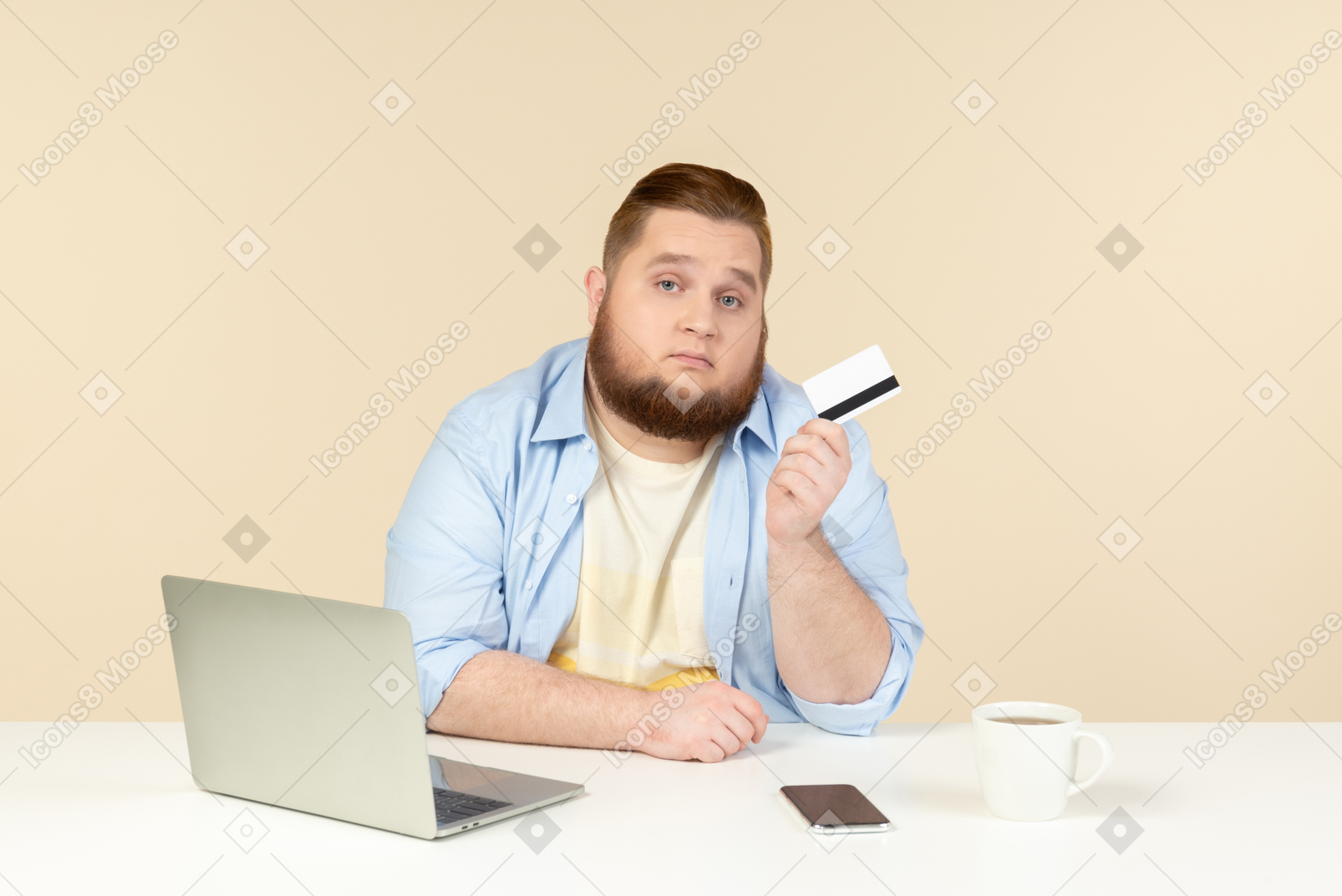 Young overweight man sitting at the table, holding phone and looking at bank card