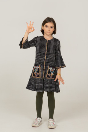 Вид спереди маленькой девочки в платье, показывающей жест ок