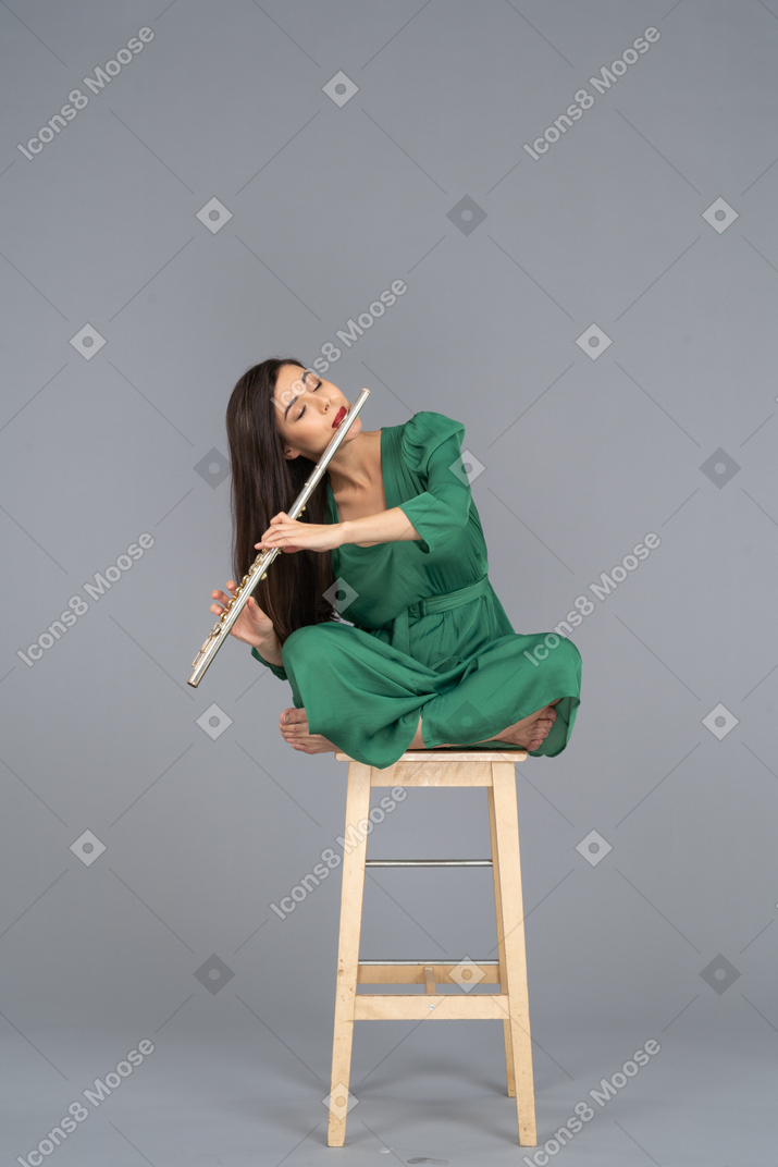 In voller länge eine junge dame, die klarinette spielt und mit gekreuzten beinen auf einem holzstuhl sitzt