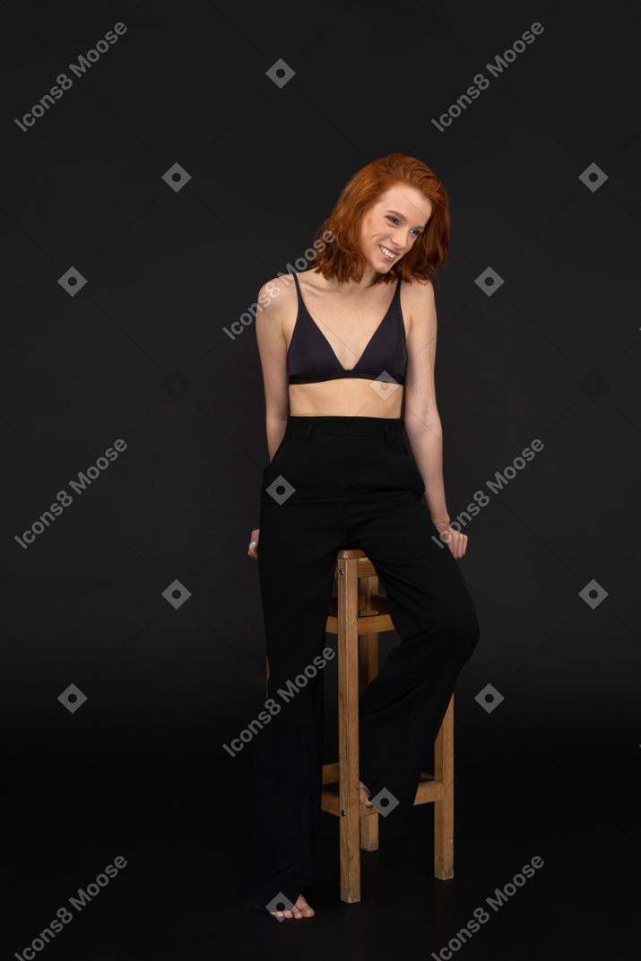 A frontal view of the sexy elegant woman, sitting on the wooden chair, smiling and looking to the right side