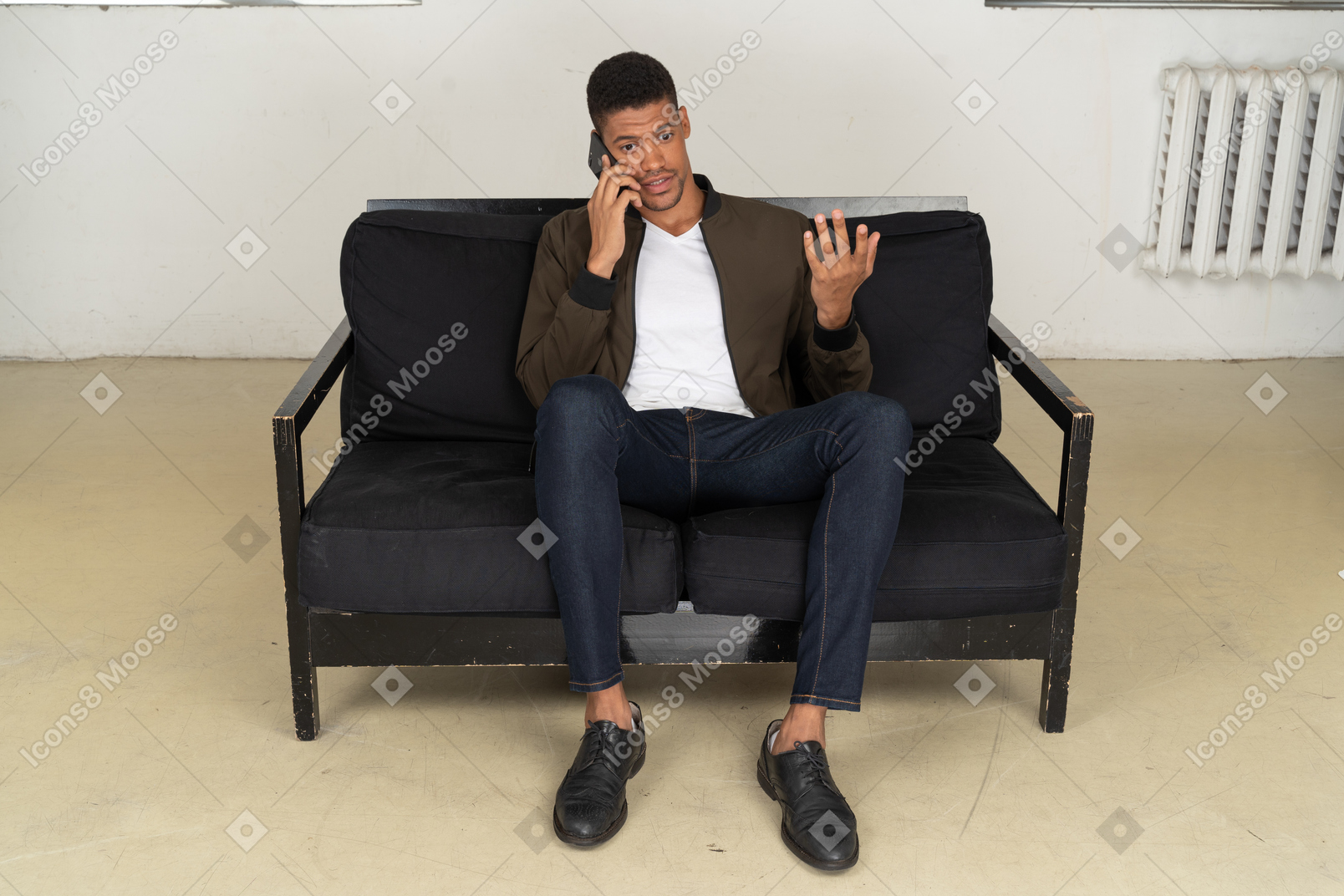 Vista frontal de un joven perplejo sentado en un sofá y hablando por su teléfono
