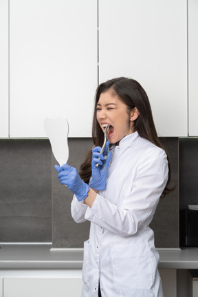 Vista de três quartos de uma médica assustada, olhando-se no espelho e tocando os dentes com um instrumento dentário