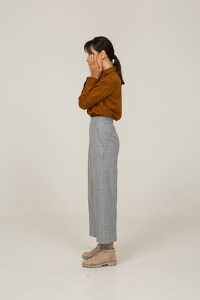 Vista lateral de uma jovem mulher asiática de calça e blusa tocando seu rosto