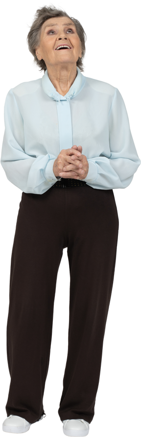 Vista frontale di una donna anziana felice in camicetta e pantaloni tenendo le mani insieme e guardando in alto
