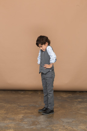 Vista lateral de um menino de terno cinza em pé com as mãos nos quadris e olhando para baixo
