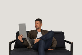 Front view of a bored young man sitting on a sofa while watching the tablet