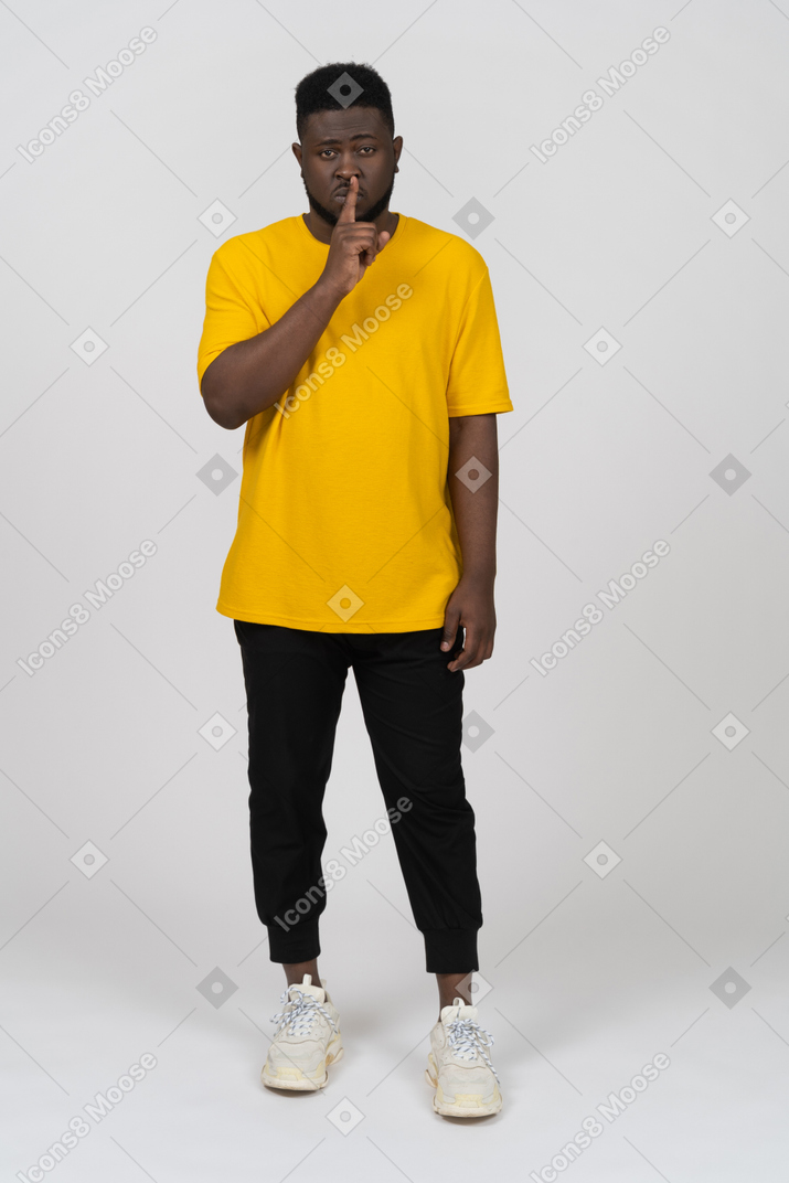 Front view of a young dark-skinned man in yellow t-shirt showing silence gesture