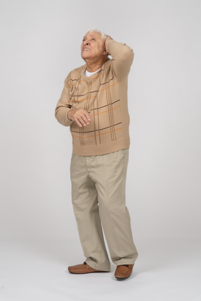 Vista frontal de un anciano con ropa informal parado con la mano detrás de la cabeza y mirando hacia arriba
