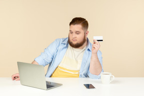 Jovem homem com excesso de peso, sentado no laptop e segurando o cartão do banco
