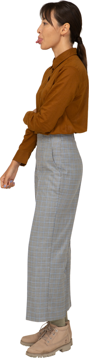 Vista laterale di una giovane donna asiatica in calzoni e camicetta che tocca il braccio e mostra la lingua