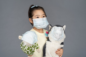 Close-up of a little girl holding two toys, all wearing face masks