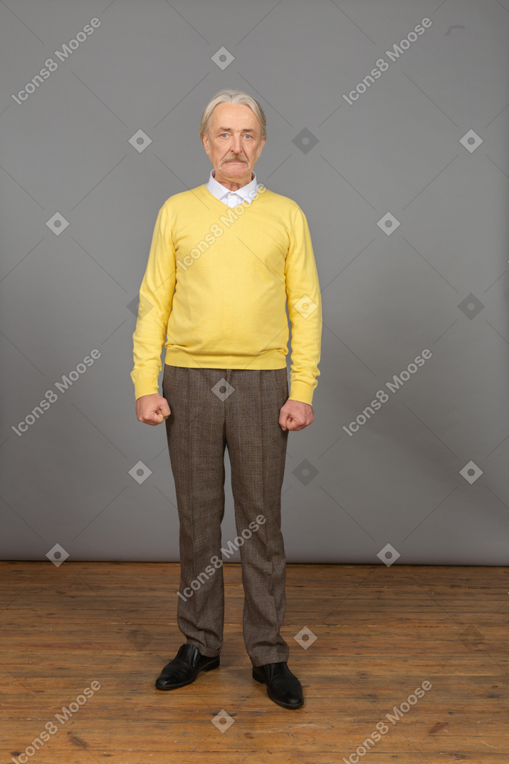 Front view of an old mad man in yellow pullover clenching fists and looking at camera