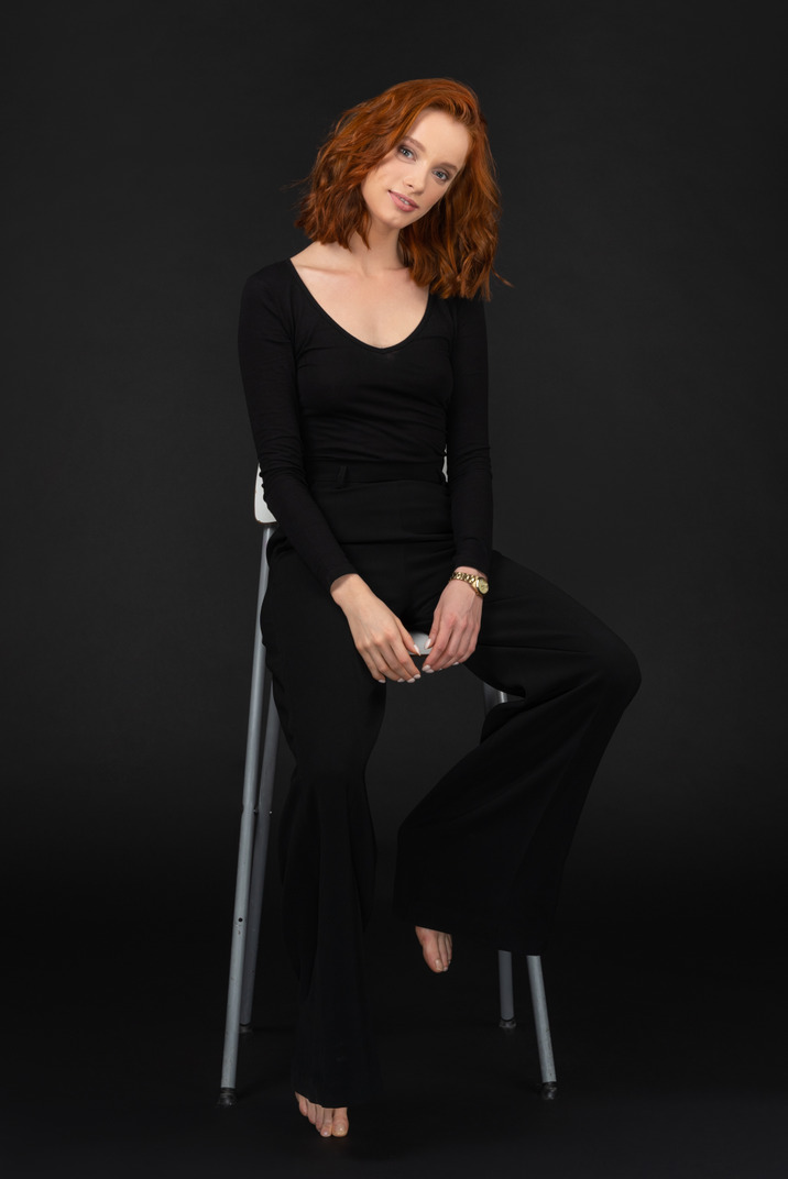 A frontal view of the cute young woman dressed in black and sitting on the tall grey chair