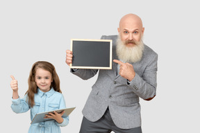 The little girl keeps the tablet and shows her finger up while the man keeps the picture and points it with a finger