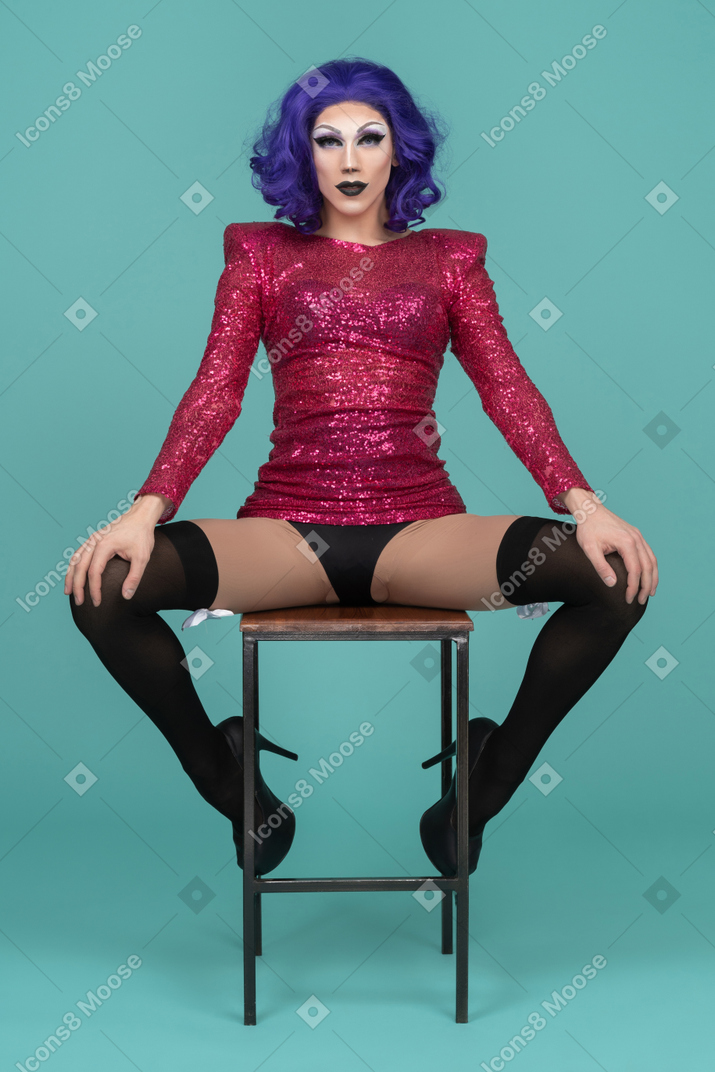Drag queen in pink sequin dress sitting on a stool with legs wide apart