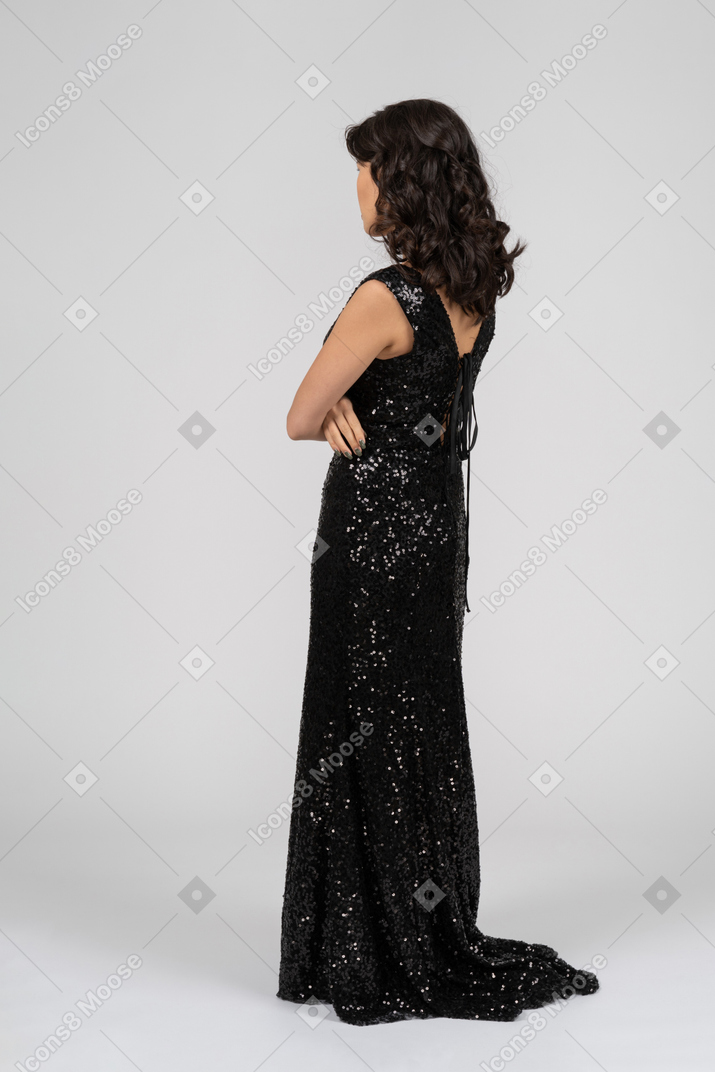 腕を組んでカメラに戻って立っている黒いイブニングドレスの女性