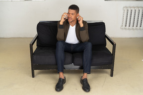 Front view of a perplexed young man sitting on a sofa and talking on his phone