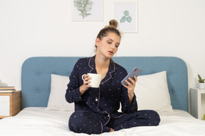 一位穿着睡衣的年轻女性坐在床上，拿着一杯咖啡，同时查看 instagram