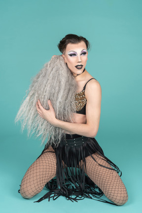 Трансвестит стоит на коленях с седым париком в руках