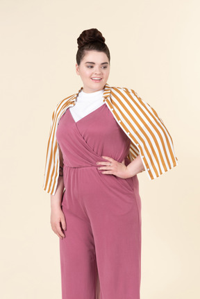 Jeune femme de taille plus dans une combinaison rose et une veste à rayures, posant sur un fond jaune pastel