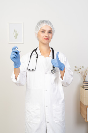 Vista frontal de una joven doctora con estetoscopio sosteniendo el termómetro y mostrando el pulgar hacia arriba