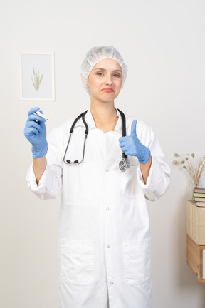 Front view of a young female doctor with stethoscope holding thermometer and showing thumb up