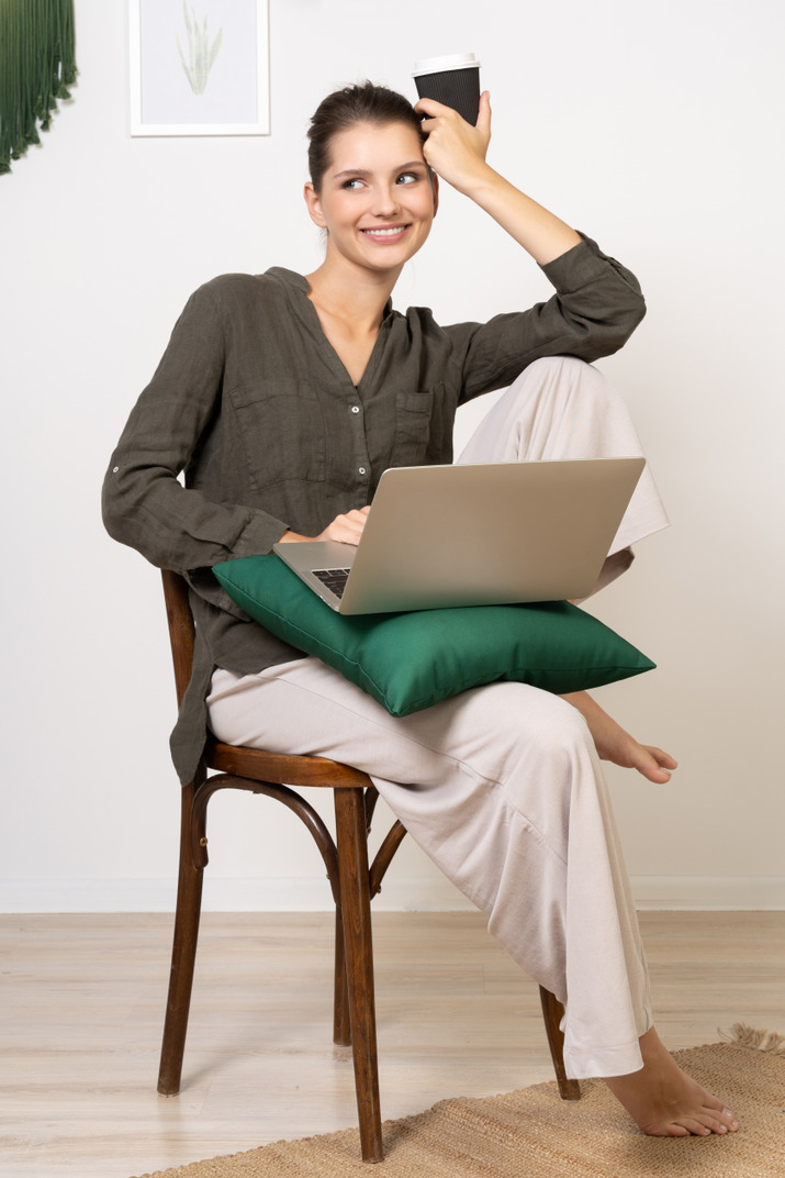 Front view of a young woman wearing home clothes sitting on a chair with a laptop and coffee