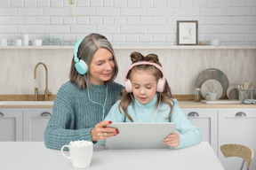 An old woman and a little girl in headphones sitting at a table with a tablet