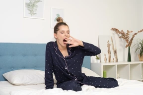 Front view of a yawning young woman in pajamas staying in bed and touching face