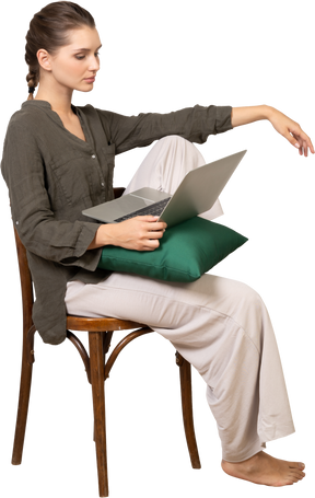 Vista laterale di una giovane donna che indossa abiti da casa seduta su una sedia con un laptop