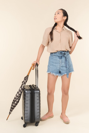 Young woman with suitcase and umbrella holding hair and looking at the sky