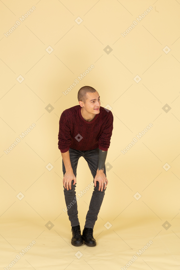 Vue de face d'un jeune homme souriant se penchant en avant et mettant les mains sur les jambes