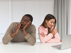 Man yawning while woman sitting at a table with a laptop