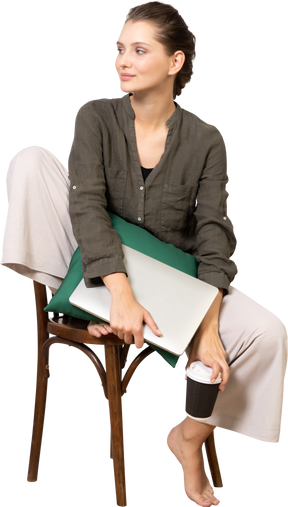 Front view of a young woman sitting on a chair and holding her laptop & touching coffee cup