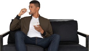 Front view of a young dreaming man sitting on a sofa while drinking coffee