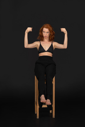 A frontal view of the confused beautiful woman dressed in black pants and bra, sitting on the wooden chair and holding her fists