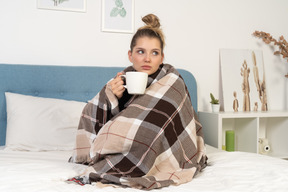 Vista frontal de una joven enferma envuelta en una manta marcada en la cama sosteniendo una taza de té