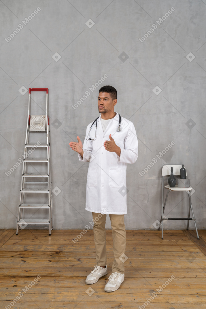 Three-quarter view of a young doctor standing in a room with ladder and chair showing a size of something