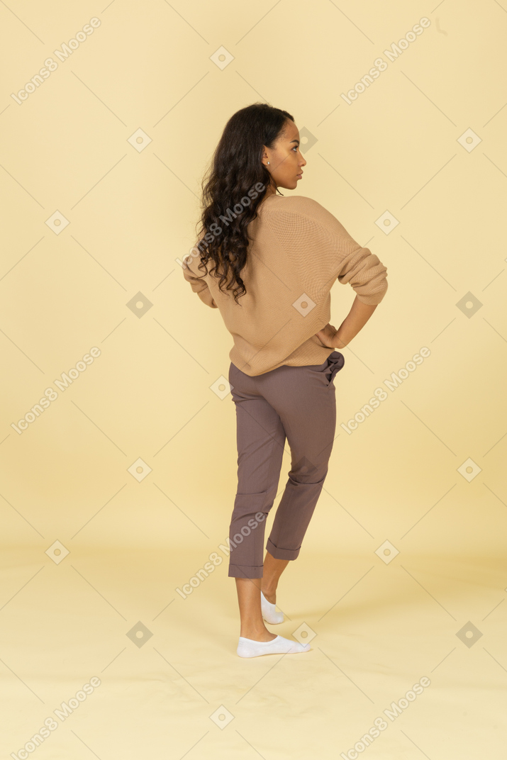 皮肤黝黑的年轻女性，将手放在臀部上的四分之三后视图